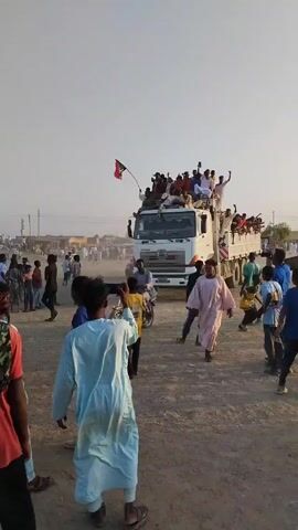 Here is a description of the video:

The video captures a celebratory scene with a large group of people gathered around a white Hino truck. Many individuals are riding on top of and in the truck's bed, waving their arms and cheering. One person is holding a flag with red, white, green, and black colors. 

The crowd is composed of people of various ages, dressed in casual clothing. They are standing and walking around the truck on a dusty ground surface. In the background, additional individuals and some buildings can be seen under a bright, clear sky. The overall atmosphere suggests a festive or rally-like event, full of energy and excitement.