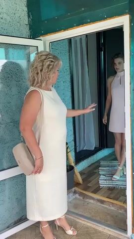 An adult woman with blonde curly hair wearing a long white dress and a beige purse on her shoulder stands outside an open doorway. She is reaching out her right hand towards a younger woman inside the doorway. The younger woman has curly brown hair and is wearing a short pink dress and white high-heeled sandals. She is reaching her right hand out towards the older woman. The older woman's left hand is by her side. The background inside the doorway shows a curtain and a wooden floor.