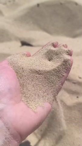 A close-up shot shows a person's hand cupped, holding a pile of light brown sand. Some of the sand is slowly trickling out from between the fingers and falling onto the sand in the background. The background consists of undulating mounds of sand.