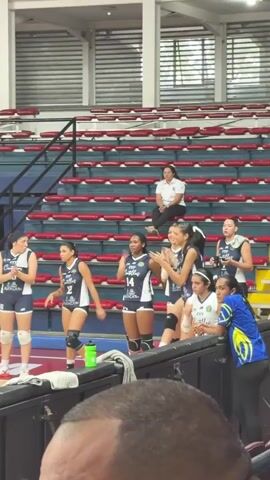 In an indoor sports arena, a group of female volleyball players wearing matching navy and white uniforms stand on the sidelines, some with their hands on the barrier, watching a game. Most of them are looking towards the court. A woman in a white top and dark pants sits alone in the mostly empty red stadium seating behind them. A green water bottle sits near the barrier. In the foreground, the top of a person’s head is visible.
