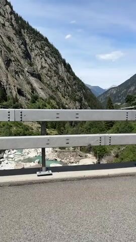 The video begins with a static shot of a mountain valley. A two-tiered metal guardrail is in the foreground, with mountains on either side. A river flows through the valley floor, its water a striking turquoise color. As the video progresses, it becomes apparent that the camera is moving, providing a panning view of the valley. The mountains are steep and rocky, with patches of green vegetation. On the right side of the valley, a covered roadway or tunnel entrance is visible, following the curve of the mountain. The sky is blue with scattered white clouds. The movement of the camera suggests it is mounted on a vehicle traveling along a road or bridge overlooking the valley. The scene is serene and captures the natural beauty of the mountainous landscape.