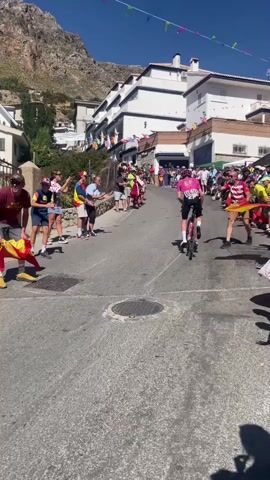 The video shows a street scene with a hill in the background. There are buildings on the right side of the street, and spectators are lining both sides of the street. A cyclist in a pink jersey with the number 203 on their back is riding up the hill. The cyclist is wearing a helmet and sunglasses. The spectators are cheering and some are holding flags. The street is paved with asphalt, and there is a manhole cover in the middle of the street. The sky is clear and blue. The overall atmosphere appears to be a sporting event, likely a cycling race.