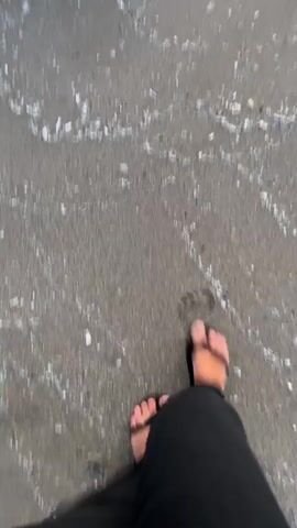 A first-person point-of-view video shows a person walking on a sandy beach. The person wears black pants and black flip-flops. Waves wash over the wet sand, covering the person's feet. Several translucent, dome-shaped objects, resembling jellyfish, are visible on the sand as the waves move back and forth.