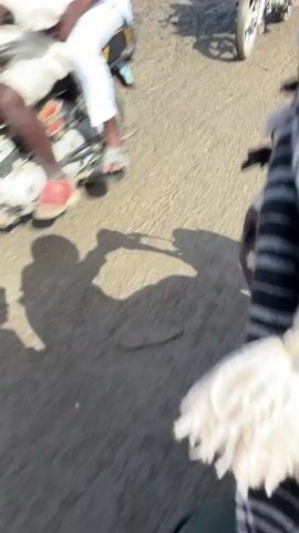 The video shows a person riding a motorcycle through a muddy, busy street, seemingly in a market area. Other motorcycles, trucks, and a yellow auto rickshaw, are also seen. The rider is wearing a black and white checkered scarf. The road is covered in mud and puddles. Shadows of the rider and the motorcycle are visible on the ground. In the background, there are buildings with red roofs and people moving around.