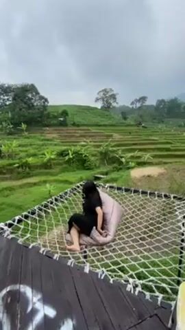 A woman in a black outfit sits on a grayish-purple beanbag chair inside a large, elevated white net. The structure with the net is made of dark wood. In the background are stepped green rice terraces under a cloudy sky. The woman adjusts her hair at one point, then looks out at the view.
