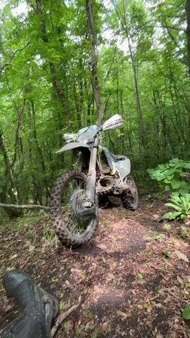 The video shows a stationary dirt bike, coated in mud, parked on a dirt path in a dense forest. The bike is positioned at a slight angle, leaning against a thin tree trunk. Green foliage surrounds the bike, with sunlight filtering through the canopy of trees. The camera slowly zooms in on the bike, revealing details such as its tires, suspension, and the number "965" on its side. A glimpse of a black motorcycle boot is visible in the bottom left corner of the frame. The overall impression is of a rugged, off-road scene.