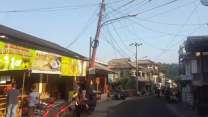The video shows a street scene in what appears to be a village or small town. On the left is a storefront with a large banner advertising "ES KELAPA MUDA HIJAU" (Green Young Coconut Ice). The storefront has wooden stalls and shelves displaying items. There are several people present, appearing to be customers or vendors. Two individuals are standing near the front, one in a blue shirt and the other in a darker shirt. A red motorcycle is parked in front of the store. A utility pole with numerous tangled wires is positioned near the storefront. Further down the street, there are several buildings, some with two stories and balconies. A paved road curves uphill. Several motorcycles are parked along the street. The scene is bathed in bright sunlight, suggesting it is daytime. The overall environment appears somewhat cluttered with overhead wires and a mix of residential and commercial structures.