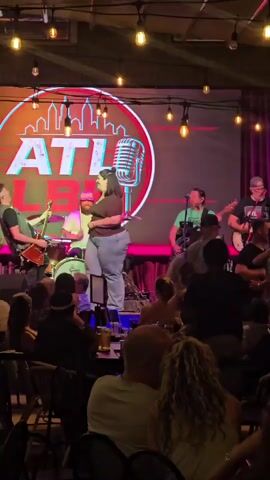 A group of musicians performs on a stage in front of a large circular screen displaying the letters "ATL" and a silhouette of a city skyline. A female vocalist, wearing a brown shirt and light blue jeans, stands center stage near a microphone. A male guitarist in a dark shirt stands to her left, playing a red electric guitar. Behind them, a drummer is seated at a drum set. Another male guitarist in a green shirt is on the right side of the stage, and another person, possibly a guitarist, in a dark shirt is to his right. The stage is lit by overhead string lights. A diverse audience is visible in the foreground, seated at tables and facing the stage. The overall atmosphere appears to be a live music performance in an indoor venue.