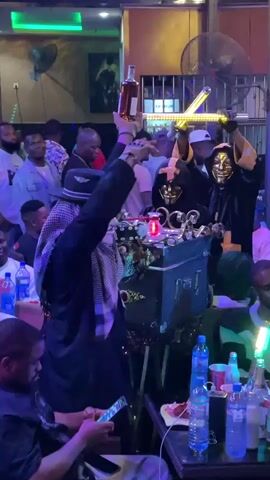 The video captures a scene inside what appears to be a bar or nightclub, where people are gathered. In the foreground, a man in a black coat and hat holds a bottle up in the air. Next to him, two figures wearing masks and dark robes hold illuminated crosses above a decorated box. Several individuals sit at tables around the room, with bottles of water and other drinks visible on the tables. The atmosphere is lively, with music playing in the background. The lighting is dim, with various colored lights adding to the ambiance of the space.