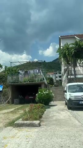 A video shows a bus stop with a map and a backpack. The camera then pans to a residential area with houses, trees, and a fence. The video continues with a shot of a house with a large hedge and a pink flower. Finally, the video shows people sitting on a porch under a wooden roof.