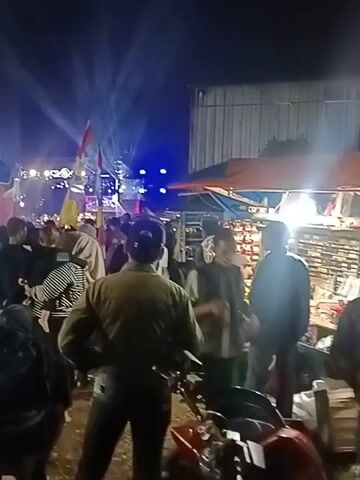 The video shows a crowded outdoor night event. There are multiple tents and stalls set up, with some featuring lights and displays. In the background, a stage area with bright lights and what appears to be sound equipment is visible. People are gathered around, some appearing to be patrons of the stalls, while others are watching the stage. A man in a green jacket is prominent in the foreground, facing away from the camera, appearing to observe the crowd. The ground is dirt or gravel. There are flags visible, including what looks like a national flag. The lighting suggests it is nighttime, with artificial lights from the stage and stalls illuminating the scene.