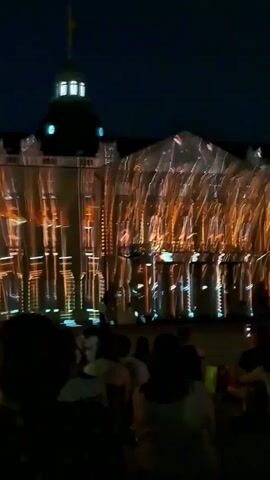 The video captures an outdoor nighttime event where a building's facade is illuminated with a projection mapping display. In the foreground, the silhouettes of a crowd of people are visible, facing the illuminated building. The building itself is adorned with dynamic, colorful light projections that create abstract geometric patterns and architectural illusions. There is a fountain in the middle ground, also partially lit by the projections. The sky above is dark.