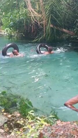Two young men are swimming in a clear, blue-green body of water, possibly a river or stream. Each is using a black inner tube as a flotation device. They are moving through the water with swimming motions. The surrounding environment is lush with green vegetation and trees, some of which overhang the water. At the edge of the water, a man is standing and watching the two swimmers. The water is clear enough to see the bottom in some areas. The overall scene appears to be one of leisure and recreation in a natural setting.