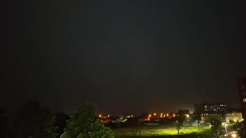 A video captures a lightning strike illuminating the night sky over a town. The sky is dark, with distant streetlights visible in the town below. Trees are silhouetted in the foreground. The lightning flash is brief and intense, casting a purple glow across the sky and momentarily revealing the details of buildings and houses. The video seems to be filmed from a balcony or window.
