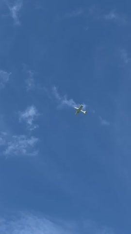 A small white airplane is captured in flight against a vivid blue sky dotted with wispy white clouds. The plane, which appears to be a single-engine propeller aircraft, is positioned in the center of the frame, slightly tilted. Its wings are extended, and the tail is visible. The overall impression is one of peaceful flight and open skies. The image seems to have been taken during daylight hours, likely mid-morning or afternoon, given the bright blue sky and the way the clouds are illuminated. There are no discernible people or text within the frame. The scene evokes a sense of freedom and the vastness of the sky. Based on the provided location context, the scene is set over Hautot-sur-Mer, France, suggesting a clear day suitable for aerial observation or recreational flying in that region.
