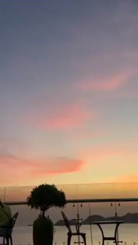 The video shows an outdoor patio scene with a view of a sunset over the water. There is a short, potted tree in the foreground, along with black metal tables and chairs. A glass barrier surrounds the patio. The sky is a blend of blue, pink, and orange, with streaks of pink clouds. A person is sitting in one of the chairs. The overall scene is serene and picturesque.