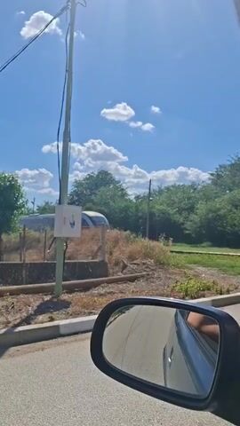 The video shows a train passing by a roadside in Bojuritsa, Bulgaria. A side-view mirror of a car is prominently featured in the foreground, reflecting the road and the sky. Beyond the car, a utility pole stands tall with a small electrical box attached to it. In the background, a train with a modern, sleek design, featuring red and white accents, travels along a railway track. The train is partially obscured by overgrown vegetation and a fence. Lush green trees and bushes line the horizon, under a clear blue sky dotted with fluffy white clouds. The sun is shining brightly, indicating it is daytime, likely late morning or early afternoon. The overall atmosphere is serene and rural, capturing a fleeting moment of movement within a peaceful landscape. No visible text is present in the video.