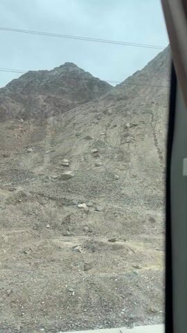 The video shows a rocky, mountainous landscape from the perspective of a moving vehicle. The scene is dominated by a large, gray-brown mountain slope covered in gravel and scattered boulders, suggesting a dry or arid environment. The sky is overcast or hazy, providing diffused light. Above the mountain, power lines are visible stretching across the frame.

On the ground near the base of the mountain, a small white sign with black Chinese characters is visible for a moment. The characters read "公路界" (Gōnglù jiè), which translates to "Highway boundary" or "Road boundary." The right side of the frame shows part of the vehicle's window frame and a beige curtain. The vehicle appears to be traveling alongside the mountain. The overall impression is one of a vast, rugged, and somewhat desolate landscape in Guazhou County, China.