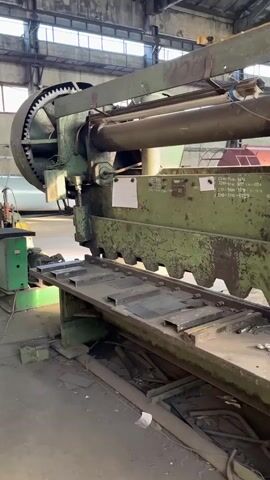 The video shows a large, industrial guillotine shear machine in a workshop. The machine is green and appears to be old and well-used, with some rust and wear visible. The machine has a long cutting blade with a serrated edge. A table in front of the blade has metal plates on it. Tools, including wrenches, are scattered on the table. In the background, there are other industrial elements, such as pipes and structural beams, and the overall environment suggests a manufacturing or metalworking facility. The camera pans slowly across the machine, giving a detailed view of its components. There are no people visible in the video.