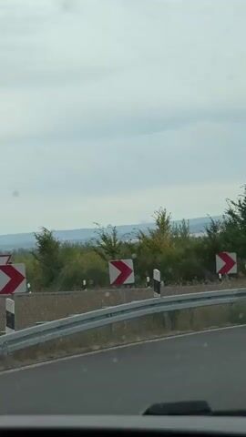 The video shows a view from inside a vehicle traveling on a road with guardrails. Several curve warning signs are visible along the roadside, indicating a series of turns ahead. The landscape consists of trees and bushes, with a cloudy sky overhead. In the distance, a truck with the logo "IMEX EXPRESS" is visible. The video is shot from a moving perspective, suggesting the vehicle is in motion.