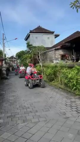 The video shows three red ATVs driving down a narrow, brick-paved street lined with buildings and foliage. The first ATV is driven by a person wearing a white shirt and red helmet, with a passenger in a tank top and red helmet sitting behind. The passenger raises their left hand in a peace sign. The second ATV is driven by a person wearing a white shirt and a red helmet. The third ATV is driven by a person in a black shirt and a red helmet. Buildings with traditional tiled roofs and a stone structure are visible along the sides of the street. Power lines run overhead against a backdrop of a light blue sky with a few wispy clouds.