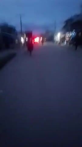 The video presents a blurry, low-light street scene at dusk or early night. The camera appears to be handheld and moving, contributing to the motion blur. A road or street extends into the distance, lined on both sides by indistinct buildings, some of which display illuminated windows or exterior lights.

The primary visible subject is a person on a motorcycle, moving away from the camera. Its prominent red taillights are the clearest feature, appearing bright against the dim surroundings. Other distant figures and vehicles are faintly discernible but heavily blurred. Utility poles with overhead lines are visible against the dark blue sky.

Artificial lights from streetlights and building interiors create a bokeh effect, with some light sources momentarily appearing as heart shapes due to optical distortion. No discernible text or specific identifying marks are visible on any objects or buildings. The overall impression is one of a busy but dimly lit street in motion, consistent with an evening setting in an urban or semi-urban area like Iwerekun, Nigeria.
