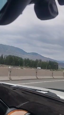 The video shows a view from the inside of a car driving on a highway. Mountains are visible in the background, with trees in the middle ground and a concrete barrier separating the lanes of the highway. The sky is cloudy.