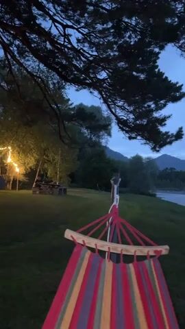 The video shows a wide shot of a serene natural landscape at dusk. In the foreground, a colorful striped hammock is suspended from what appears to be a wooden frame, its red, yellow, green, and purple fabric filling the bottom portion of the frame. To the left, a portion of a large tree with dark green needles extends into the frame from the top, its branches casting shadows. Behind the hammock, a grassy slope leads down to a wide river. The river is calm and reflects the muted, cloudy sky. On the opposite bank of the river, a dense line of dark green trees forms a natural border. Beyond the trees, rolling hills and mountains are visible in the distance, fading into the twilight haze. The sky is overcast with a soft, diffused light, indicating either early morning or late evening. There are no people or animals visible in the video. The overall atmosphere is peaceful and tranquil.
