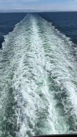 The video shows a view from a moving vessel, looking backward at the turbulent wake it is creating in the ocean. The water is a deep blue, and the sky above is a clear blue with scattered white clouds. The wake is a wide, frothy trail of white foam and churning water that extends far behind the vessel, gradually dissipating into the calmer sea. The horizon is visible in the distance, with a faint line of land or other vessels. The camera is positioned at a slight downward angle, emphasizing the vastness of the ocean and the power of the vessel.