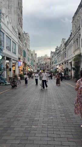 A young couple walks hand-in-hand down a cobblestone street lined with multi-story buildings. The woman, in a grey top and white skirt, looks at the man, who is wearing a white t-shirt and black pants. They stop and look up at something off-camera. Other pedestrians are visible in the background, as are shops and decorative elements on the buildings. The sky is overcast.