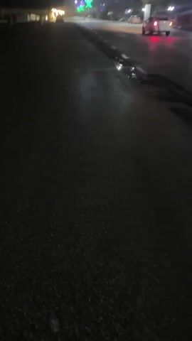 The video shows a street at night. The road is wet and reflects the light from streetlamps and car headlights. There are several vehicles on the road, including cars and possibly a bus. The lighting is dim and there appears to be fog or mist in the air, blurring the lights. The road has some potholes and cracks. There are trees lining the side of the road. There are also decorative lights in the distance, possibly for a holiday or event. The video is shaky and taken from a low angle.