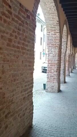 The video shows a POV shot walking along a covered walkway with brick arches. The walkway has a stone-paved floor. On the left, a white building with several windows and arched doorways is visible. A green car is parked on the cobblestone street in front of the building. The walkway continues into the distance, with more brick arches and a dark ceiling overhead. The overall environment appears to be an old European town or city.