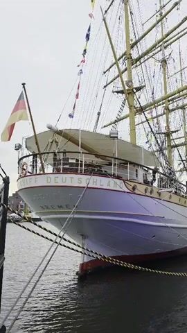 The video shows a large sailing ship named "Deutschland" docked in a harbor. The ship is white with red and gold accents and has multiple masts and sails. A German flag flies from the bow. People are visible walking on the dock alongside the ship, and there are buildings in the background. The water is dark, and the sky is overcast.