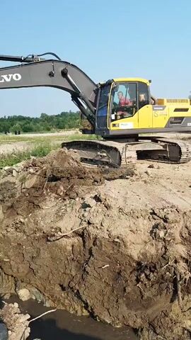 An excavator is being used to dig a trench next to a body of water. The excavator is yellow and black, with a Volvo logo on its arm. There are two people inside the cab of the excavator, both wearing red shirts. The excavator is digging up a large amount of dirt and debris, which is being piled up next to the trench. The trench is partially filled with water. The surrounding environment is a rural area with trees and fields in the background. The sky is clear and blue.