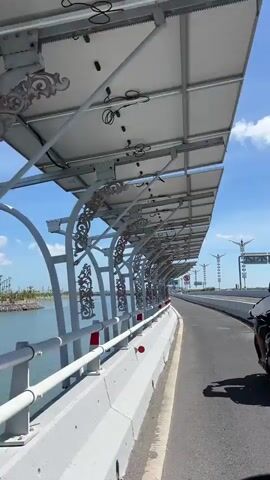 The video is a first-person perspective from a moving vehicle, likely a motorcycle, on a sunny day on a bridge. To the left, there is a body of water. The bridge has a white railing with red and white stripes. Above the railing, there are decorative metal panels. The bridge is covered by a structure with many solar panels on top, supported by white metal beams and arches. A motorcycle is visible ahead on the right side of the road. Traffic signs are also visible on the right side of the road. The sky is blue with some scattered clouds. The video captures the view as the vehicle travels across the bridge.