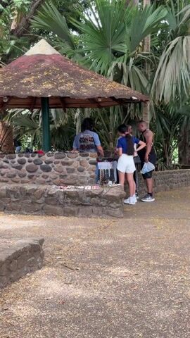 An outdoor vendor stall with a rustic stone counter and a conical thatched roof is visible under bright daylight, surrounded by lush tropical foliage, including large palm trees.

At the center of the image, a vendor, a man with long dark hair wearing a blue t-shirt, stands behind the stone counter, attending to the merchandise. To his right, three individuals are browsing items. A woman with long dark hair tied in a ponytail (with a pink hair tie) wears a blue short-sleeved top, white shorts, and white sneakers, leaning over the counter to examine products. Behind her, another woman with lighter hair and top is partially visible. To the far right, a man with short dark hair, wearing a black sleeveless top, black shorts, and black sneakers, stands observing the items. He has a tattoo on his right arm and is holding a black plastic bag in his left hand.

The stall displays various items for sale, primarily bracelets and necklaces, hung on racks and laid out on the stone counter. Some items on the left side of the counter feature red, black, and white patterns. The ground is a dirt path scattered with small yellow leaves or petals. In the background to the left, under the same thatched structure, another person is sitting on a low stone wall. The overall scene suggests a casual market or tourist area in a warm, outdoor environment.