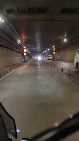 A first-person perspective video shows a view from a vehicle traveling through a dimly lit tunnel. The tunnel is lined with lights and has reflective markings on the walls. Orange traffic cones are spaced along the right side of the road. In the distance, headlights from other vehicles can be seen. The road surface appears wet, with reflections of the lights visible. A white SUV is parked on the right side of the tunnel ahead.