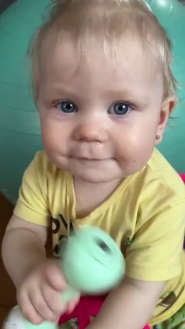 A baby with blond hair and blue eyes sits and looks at the camera. The baby wears a yellow t-shirt with text and a cartoon animal on the front. The baby has a pink toy steering wheel in front of them and holds a green toy in their left hand. The baby's face is dirty. A green exercise ball is behind the baby. The baby makes a face and then appears to cry.