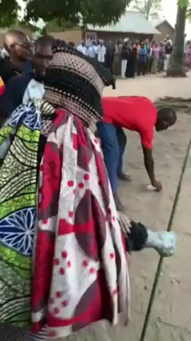 The video shows a crowd of people gathered in an outdoor setting. The focus is on a person in the center, heavily adorned in layered, colorful garments, and a large, possibly woven, hat that obscures their face. This person appears to be dancing or moving in a rhythmic manner. Around them, other people are moving as well, with some recording the event on their phones and others watching. The environment appears to be a village or rural area, indicated by the presence of simple buildings in the background and the sandy ground. The overall atmosphere seems festive and communal.
