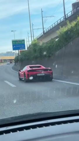 From inside a moving car, the camera records a red Ferrari driving in the same direction. The Ferrari is in the lane to the right, and a metal bridge is visible above. Road signs indicating directions to Opfikon and Glattbrugg are visible to the left of the screen. The Ferrari has a Swiss license plate.