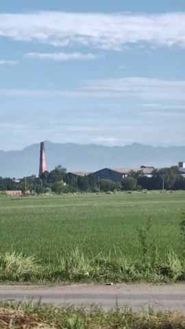A video shows a landscape with a brick chimney and a factory in the background. A green field with some people is in the foreground, and a statue of Buddha is on the right side of the screen. The sky is blue with some clouds. The video appears to be shot from a moving vehicle.