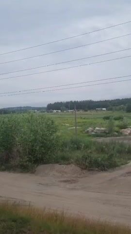 The video shows a rural landscape under a cloudy sky. A dirt road runs along the bottom of the frame. On the left side of the road, there is tall grass and some bushes. In the middle ground, there is a large, open field with patches of green grass, some weeds, and scattered bushes. In the distance, there is a line of trees and some buildings. Power lines stretch across the top of the frame. A red SUV with decals drives down the dirt road from left to right. A utility pole with insulators and wires stands in the field.