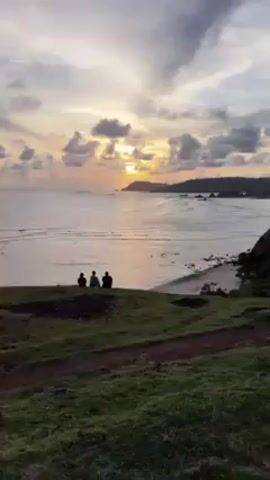 The video shows three people sitting on a grassy hill overlooking the ocean at sunset. The sun is partially obscured by clouds, casting a warm glow on the water. The beach is visible in the distance, with waves breaking on the shore. The silhouettes of the people on the hill create a peaceful and contemplative scene.