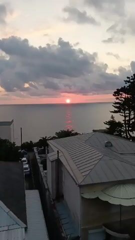 The video is a vertical shot of a sunset over the ocean. The sun is a bright, round orb low on the horizon, casting a shimmering pink reflection on the water. The sky is filled with dramatic, dark clouds tinged with pink and purple hues. In the foreground, the roof of a white building with a gray, corrugated roof is visible. To the left of the building, a dark fence and some parked cars can be seen. Palm trees are also visible near the building. The scene is calm and serene, with the focus entirely on the natural beauty of the sunset.