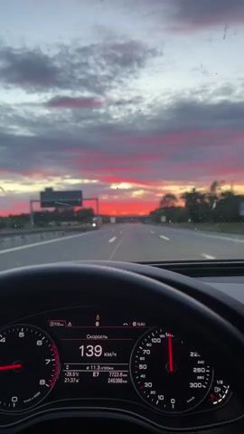 The video shows a point-of-view shot from inside a car driving on a highway at dusk. The dashboard displays a speed of 137 km/h, an outside temperature of 11.2 degrees Celsius, and a distance of 7700 km and 24533.8 km. The sky is filled with dramatic red and pink clouds, indicating a sunset. The highway has multiple lanes and is relatively clear, with a few other vehicles visible in the distance. An overpass is visible ahead. The driver's hands are on the steering wheel.