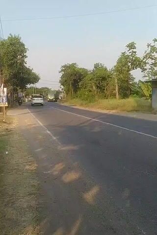The video shows a roadside scene. On the left side of the road, there is a wooden pole with a sign, a red motorcycle, and a white car. Further down the road, a large green truck is visible. On the right side of the road, there is a ditch with dry grass and trees. The road is paved and appears to be a two-lane highway. The sky is clear and blue. There are some shadows on the road, suggesting it is daytime. In the foreground, the camera is positioned close to the edge of the road, with a grassy area on the left. As the video progresses, several vehicles pass by: a red SUV, a motorcycle with two occupants, and another motorcycle. The camera appears to be moving, following the flow of traffic. The overall impression is of a rural or semi-rural setting with moderate traffic.