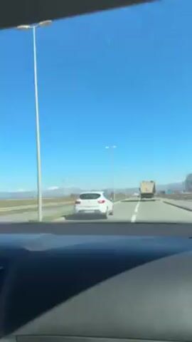 From the interior of a moving car, the video depicts a clear, sunny day on a highway. A white car is in the same lane ahead, followed by a truck further in the distance. The road is bordered by greenery on one side and the horizon shows distant, snow-capped mountains. Tall streetlights line the highway. The camera remains static within the vehicle, capturing the forward view of the road.