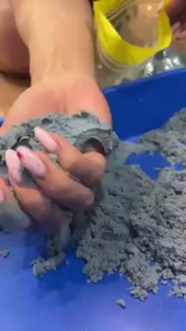 A close-up video shows a person holding gray kinetic sand in their hand. The person has pink-painted fingernails with white tips. The sand is crumbly and textured. Below the hand is a blue plastic tray. A yellow container is visible in the background, partially out of focus. The background is a wooden floor. The person is holding the sand in a cupped hand, and some sand is falling back into the tray.