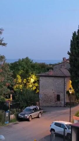 The video shows a view of a small Italian village at dusk. The sky is a pale blue, transitioning to a soft pink near the horizon. In the foreground, a paved road curves through the scene. On the left side of the road, a silver pickup truck is parked. To the right, a white SUV is parked facing the viewer. Behind the SUV, a small red-roofed building is partially visible.

The middle ground features traditional stone buildings with terracotta roofs. One building has a lit window, casting a warm glow. Several tall cypress trees stand prominently in the center of the frame, their dark green foliage contrasting with the muted tones of the buildings and sky. In the background, more buildings, including a church tower with a bell, are nestled among trees on a hillside. Lush green vegetation covers the slopes. The overall atmosphere is serene and peaceful, characteristic of a quiet evening in the countryside.