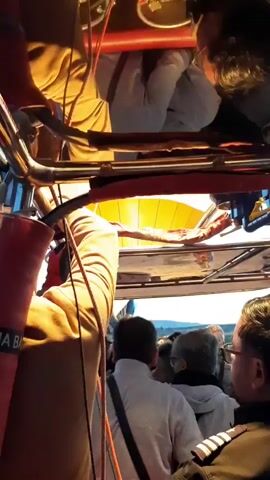 This is a video taken from inside a hot air balloon basket. People are visible in the basket, some looking out at the scenery. The video pans to show another hot air balloon in the distance, which is inflated and preparing for flight on a grassy field with some vehicles nearby. The sky is a clear blue, and mountains are visible in the background. The overall impression is of a sunrise or early morning hot air balloon launch.